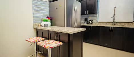 A kitchen with a stainless steel refrigerator, dark cabinets, a granite countertop, and two bar stools with patterned cushions.