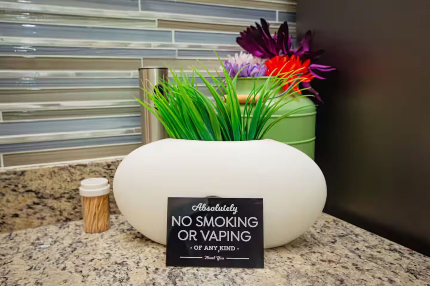 Un vase blanc au design moderne, posé sur un comptoir de cuisine, sur lequel est apposé un panneau indiquant : « DÉFENSE DE FUMER OU D’UTILISER DES CIGARETTES ÉLECTRONIQUES ».