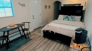A bedroom with a bed, a desk, a chair, a coffee maker, and a window with blinds.