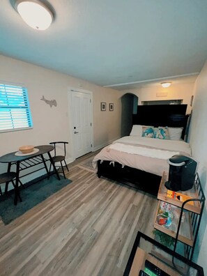 Un dormitorio con cama, escritorio, silla, cafetera y ventana con persianas.