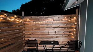 A patio with a wooden wall, two chairs, a table, and string lights.