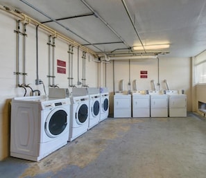 A laundry facility with multiple washing machines and dryers, pipes on the ceiling, and warning signs on the walls. - New Wonderful Beach Front Studio in Daytona Beach! (Daytona Beach)