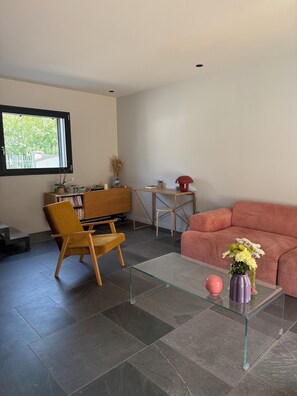 A modern living room with a glass coffee table, a sofa, a wooden chair, a small side table, and a window with a view of greenery. - A charming house in Petit Port, between the lake and the mountains (Annecy)