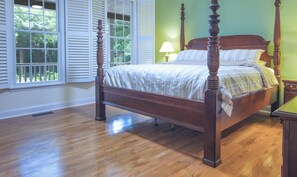 Uma cama de dossel com estrutura de madeira e colcha branca, em um quarto com piso de madeira e vista para o verde através de grandes janelas com persianas brancas.