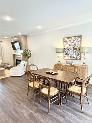 Un coin repas comprenant une table en bois, six chaises, une cheminée, un téléviseur fixé au mur et un tableau décoratif.
