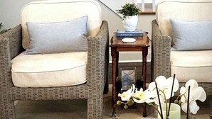 Una habitació amb dues cadires de vimet, una taula auxiliar de fusta, un llibre i un ram de flors blanques.
