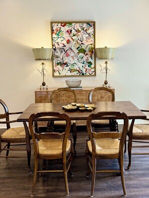 A dining room with a wooden table, six chairs, two wall-mounted lamps, and a large abstract painting. - Meet Me In Oxford- 3BR -1 Mile from the Square (Oxford)