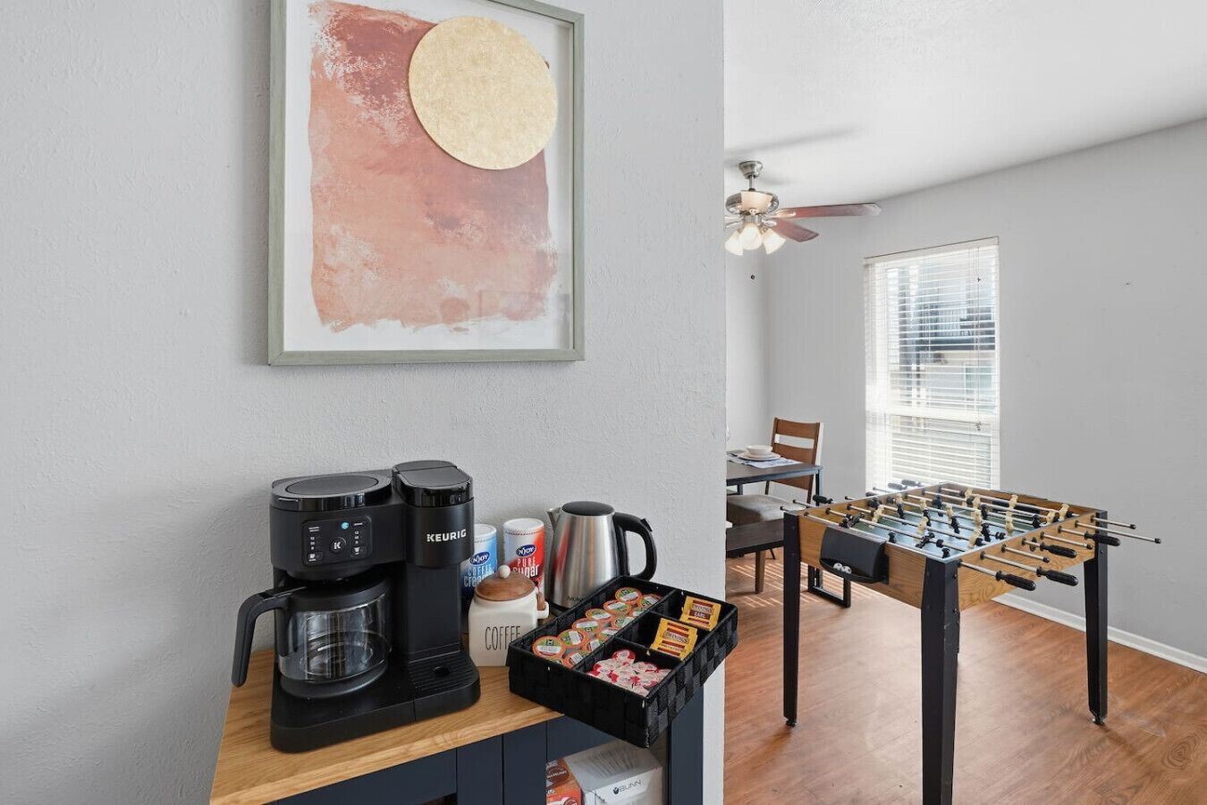 Een kitchenette met koffiezetapparaat, waterkoker en koffiekopjes, een tafelvoetbaltafel en een ingelijst abstract schilderij.