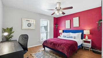 A bedroom with a bed, a desk, a chair, a ceiling fan, and a window.