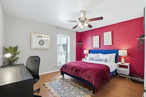 A bedroom with a bed, a desk, a chair, a ceiling fan, and a window.