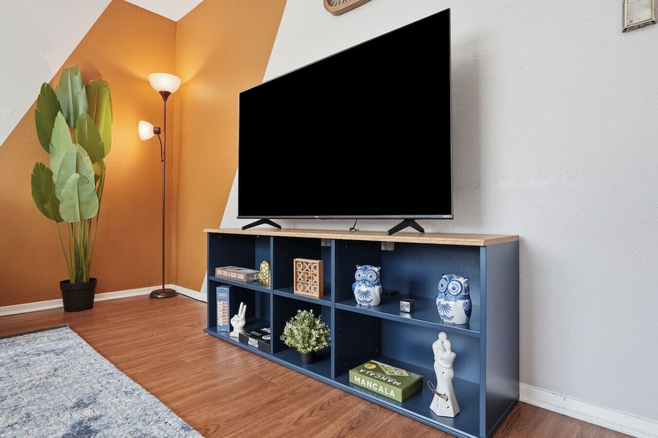 A living room with a flat-screen TV mounted on a wooden stand, a potted plant, and a blue shelving unit with decorative items.