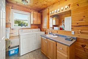 A compact kitchen with wooden cabinets, a built-in washer and dryer, a sink, and a window with a view of greenery.