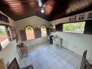 A room with a tiled floor, wooden ceiling, and a desk with a chair. There are windows with shutters, a radiator, and framed pictures on the wall. - Beach house with pool in Porto das Dunas, 8 suites, 5 min from the beach. (Aquiraz)