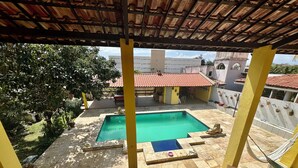A pool area with a tiled deck, a covered seating area, and a view of a building with a red-tiled roof. - Beach house with pool in Porto das Dunas, 8 suites, 5 min from the beach. (Aquiraz)
