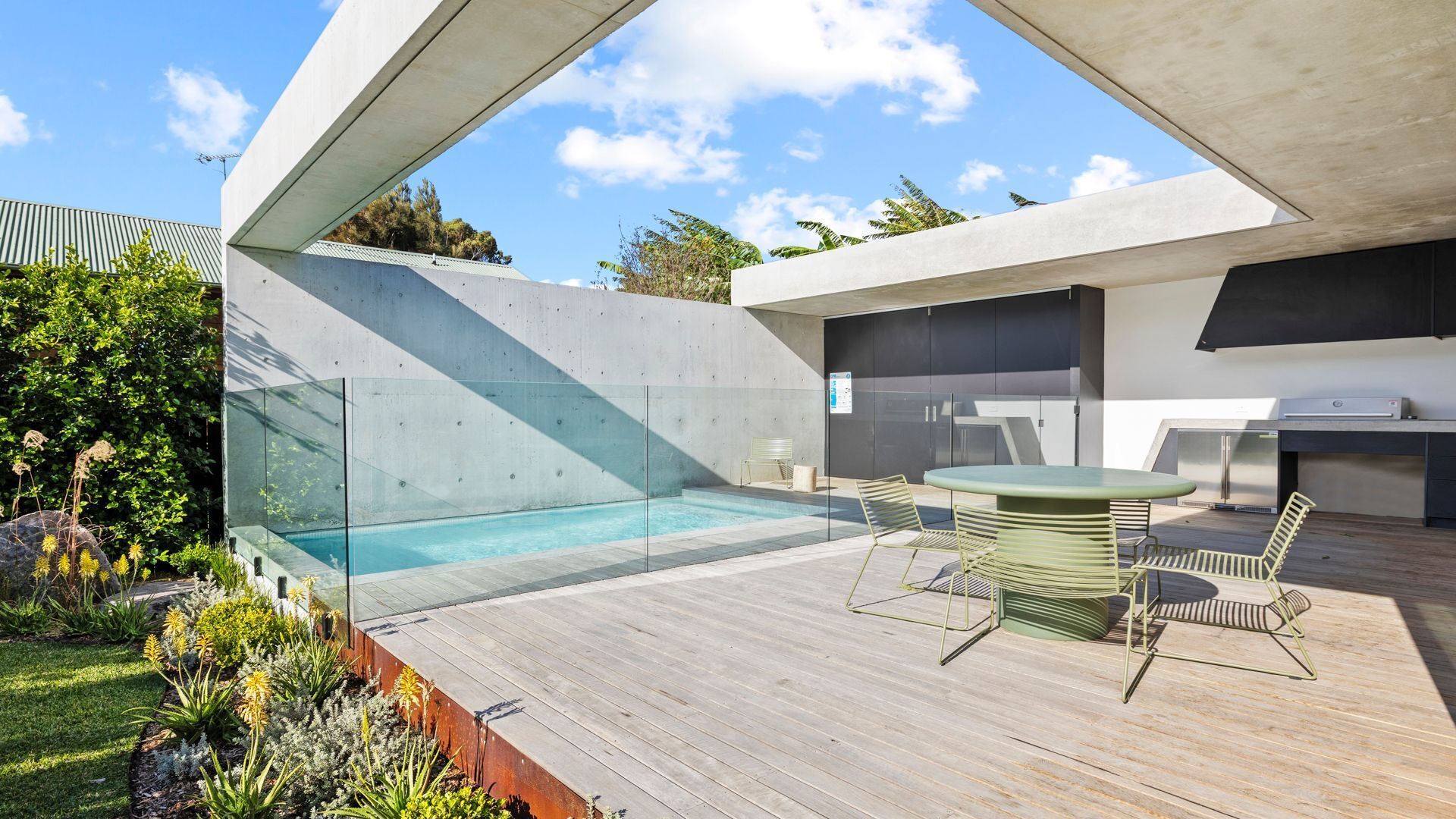Area piscina esterna moderna con recinto in vetro, terrazza in legno e barbecue integrato.