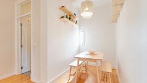Una habitación con piso de madera, una mesa blanca con dos sillas, una lámpara colgante y un estante de madera con adornos.