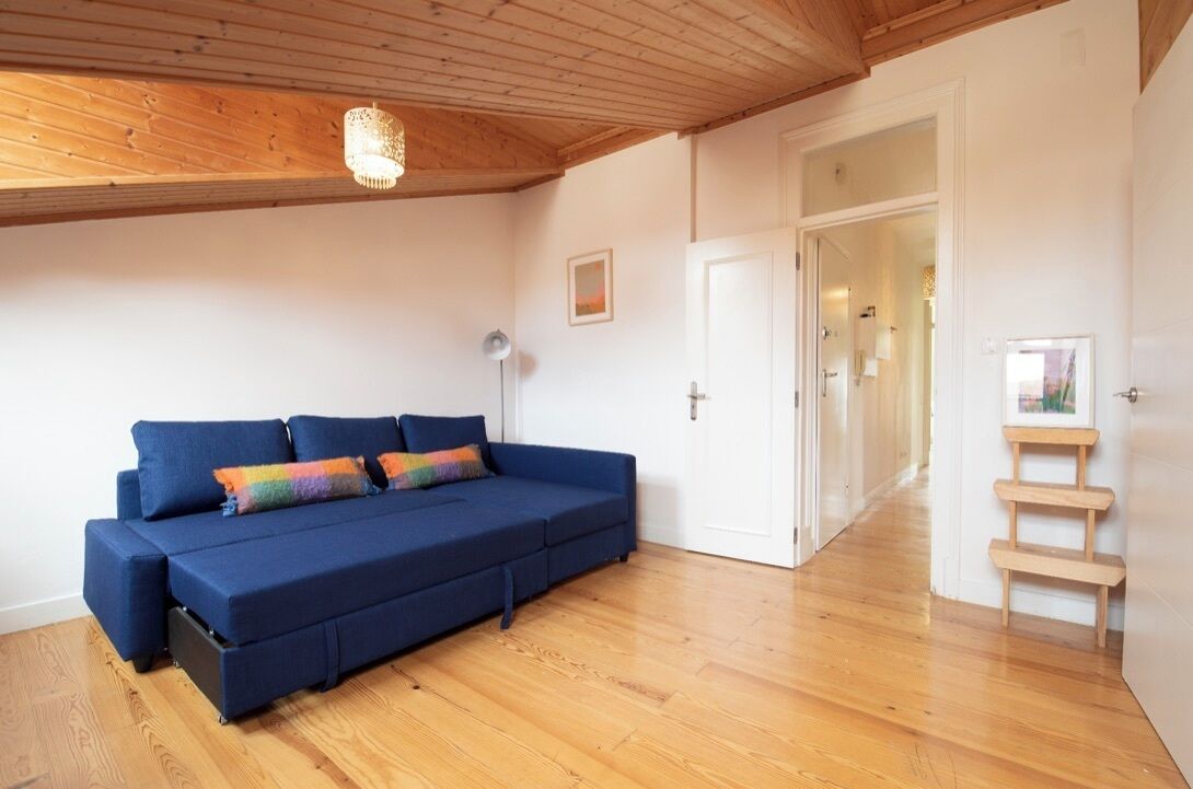 A room with a blue sofa, wooden flooring, a ceiling light fixture, and a door leading to another room.