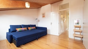 Una habitación con un sofá azul, suelo de madera, una lámpara en el techo y una puerta que da a otra habitación.