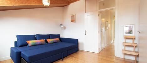 A room with a blue sofa, wooden flooring, a ceiling light fixture, and a door leading to another room.