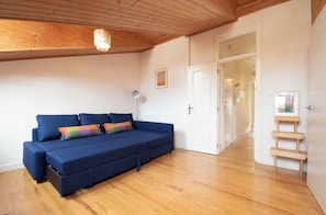 Ein Zimmer mit blauer Couch, Holzboden, einer Deckenleuchte und einer Tür zu einem anderen Raum.