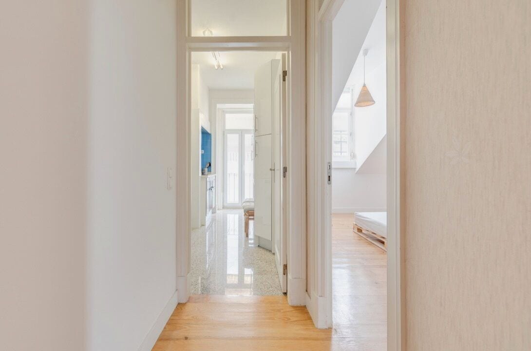 A hallway with a wooden floor leading to a room with a bed and a hanging light fixture.