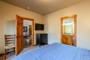 Ein Schlafzimmer mit einem Bett, einem Holzstuhl, einer Kommode, einem Fernseher und einem Badezimmerspiegel.