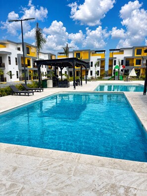 Una piscina con sillas de descanso, una zona de estar techada y un edificio con paredes de color amarillo y blanco.