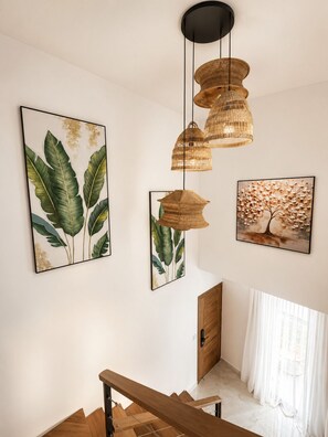 Eine Treppe mit Holzdielen und Handlauf, eine Wand mit drei gerahmten Pflanzenbildern und einer hängenden gewebten Leuchte.