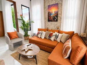 Ein Wohnzimmer mit Sofa, Sessel, Couchtisch und einem großen Fenster mit Vorhängen.