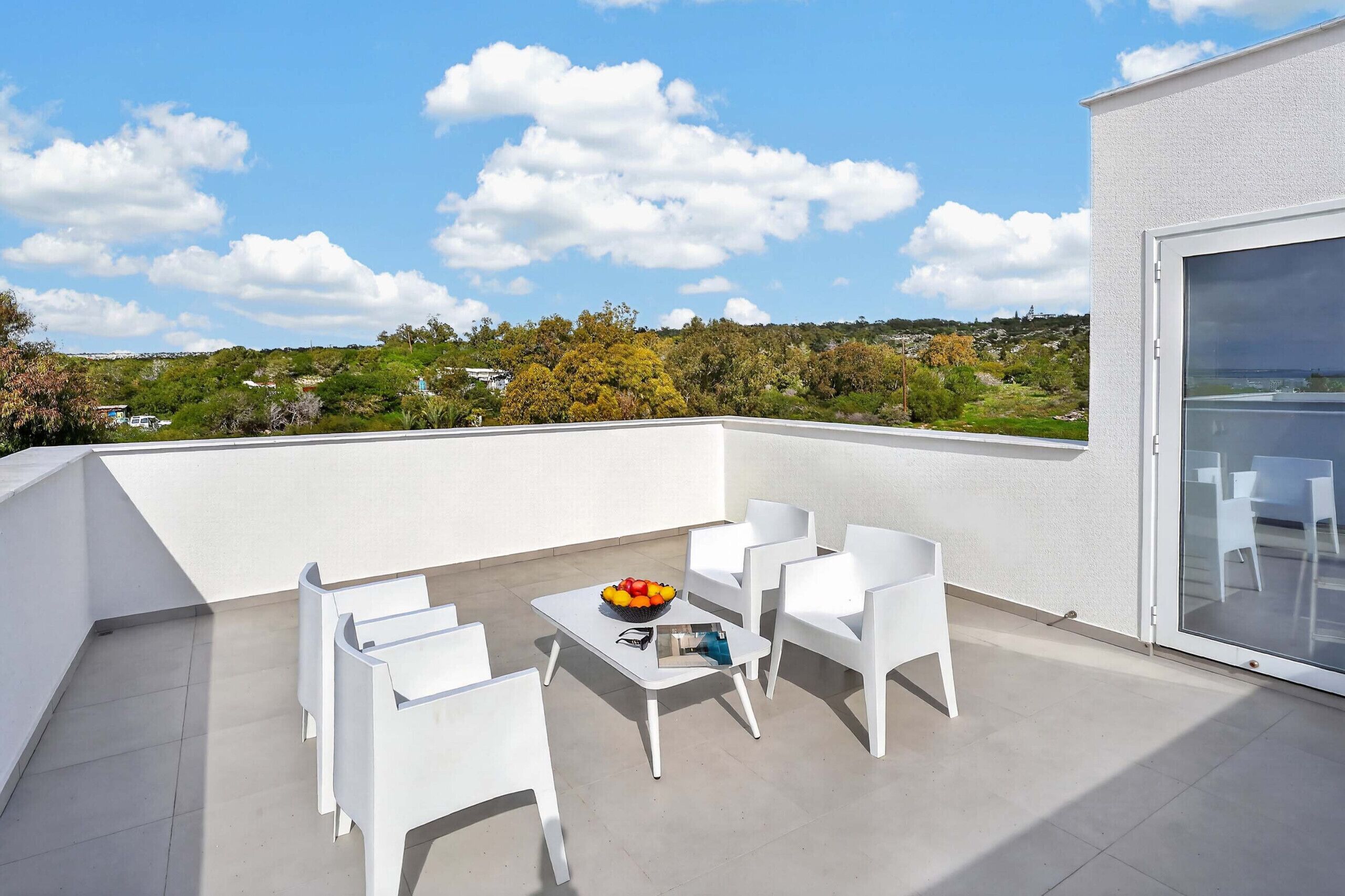 A rooftop terrace with white chairs and a table, a bowl of fruit, and a view of a hilly landscape.