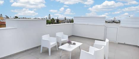 A rooftop terrace with white chairs and a table, a clear blue sky, and a view of the cityscape.