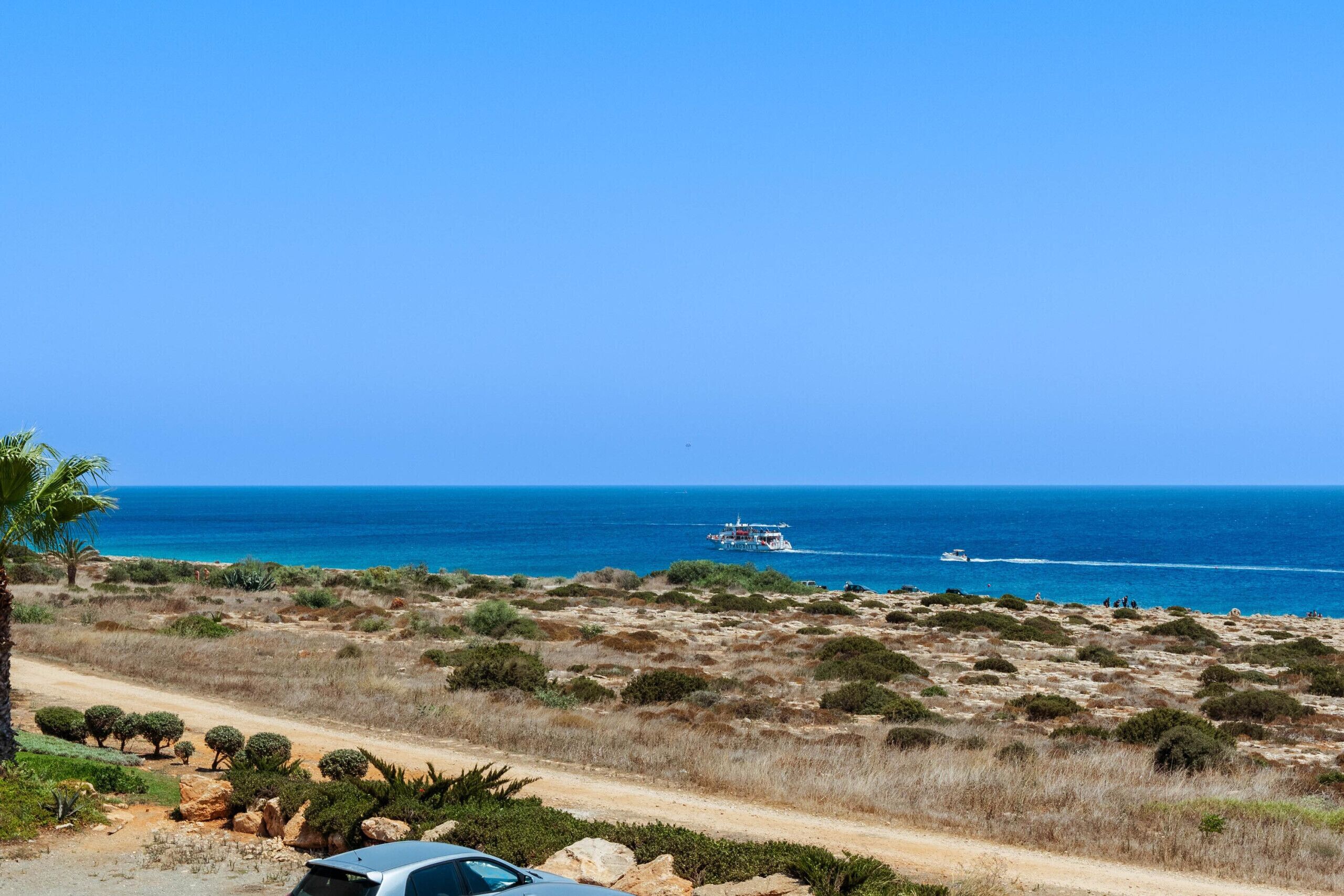 A coastal road with sparse vegetation, a clear blue sky, and the sea in the background.