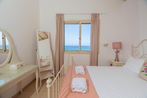 A bedroom with a bed, a bedside table with a lamp, a dresser with a mirror, a window with curtains, and a view of the sea. - Artisan Seafront Villa - Escape (Protaras)