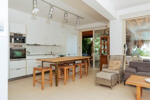A modern kitchen with a wooden table and bar stools, a dining area with a sofa and chairs, and a glass door leading to an outdoor area. - Artisan Seafront Villa - Treehouse (Protaras)