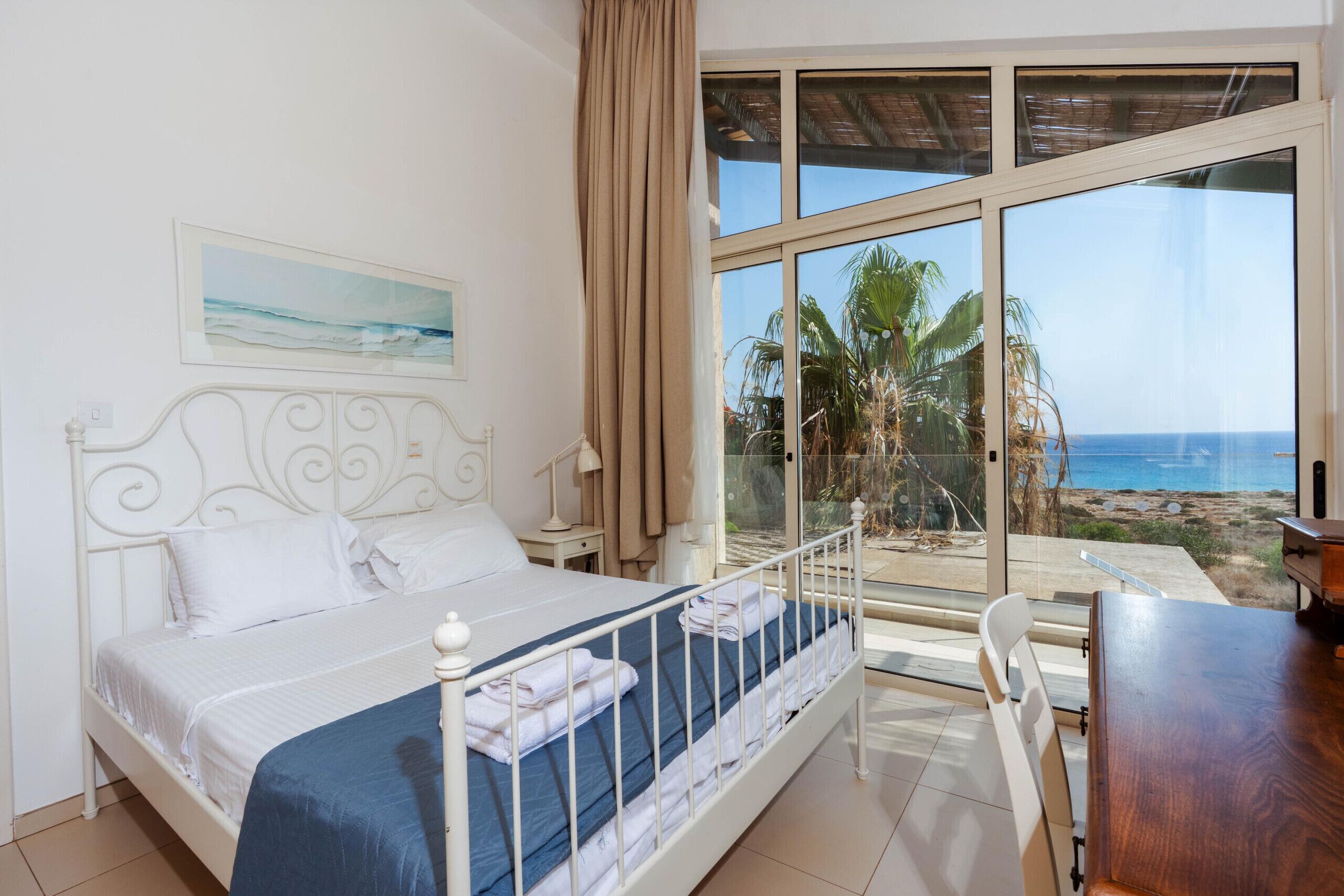 A bedroom with a bed, a nightstand, a lamp, a window with a view of the beach and palm trees.