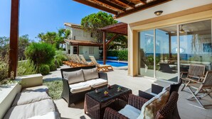 A covered outdoor seating area with wicker furniture, a pool, and a building with a tiled roof in the background. - Artisan Seafront Villa - Treehouse (Protaras)