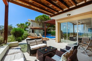 A covered outdoor seating area with wicker furniture, a pool, and a building with a tiled roof in the background. - Artisan Seafront Villa - Treehouse (Protaras)