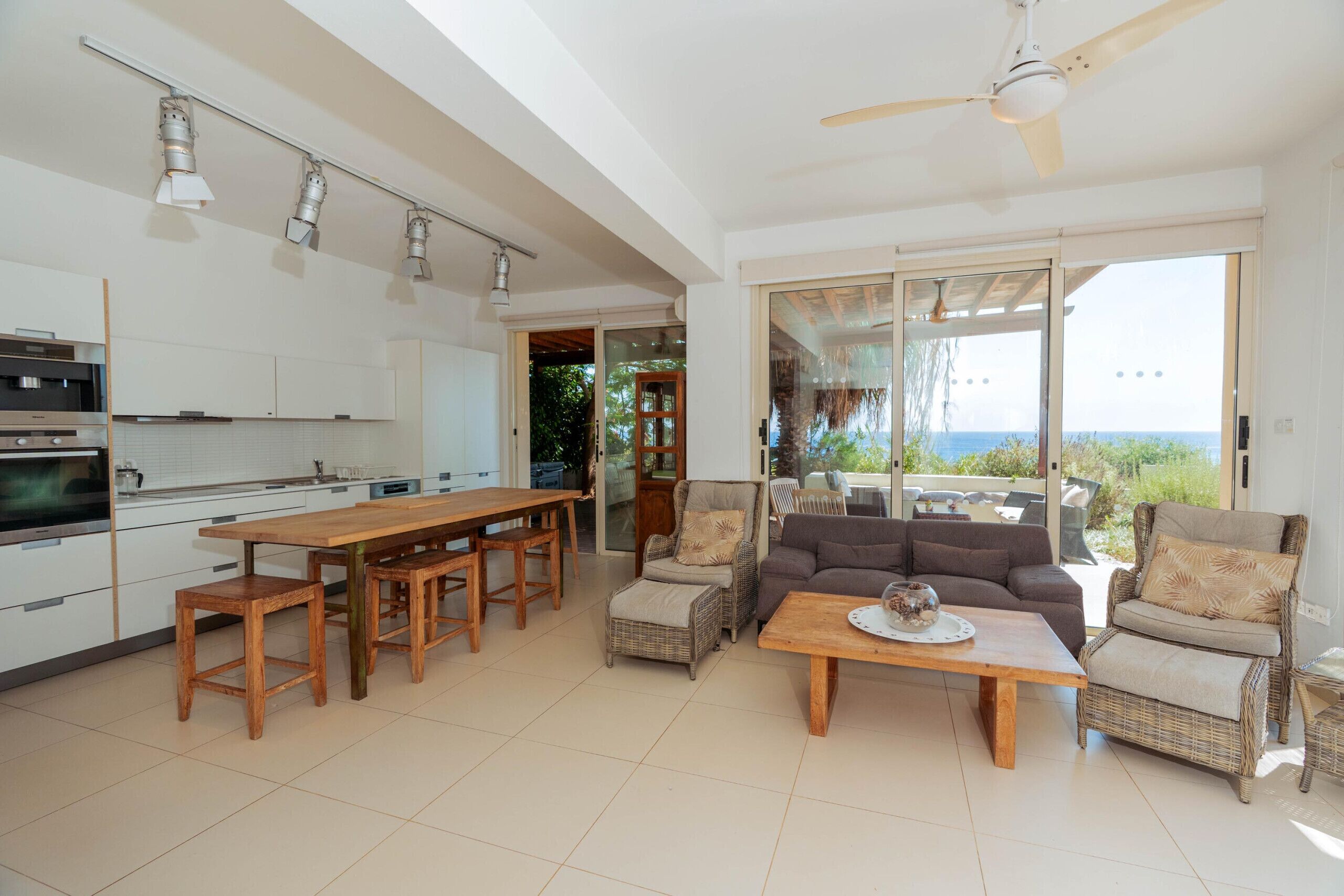 A modern kitchen with a dining area, wicker furniture, and a view through large glass doors.