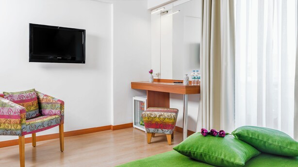 Ett hotellrum med en tv som hänger på väggen, ett skrivbord i trä med en spegel, en färgglad stol, en grön soffa med kuddar och ett litet kylskåp.