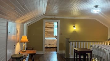 Une pièce avec un plafond en bois, un lit recouvert d’une courtepointe blanche, une table avec une lampe et une table à manger avec des magazines.