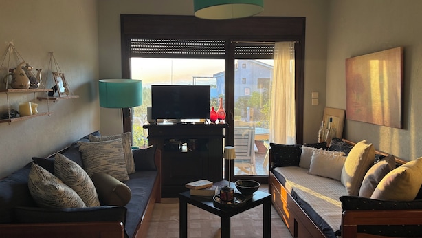 A living room with a sofa, a coffee table, a TV, and a large window with a view. - Halkidikigr (Nikiti)
