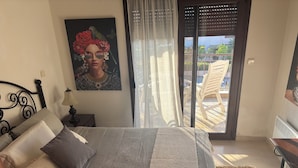 A bedroom with a bed, a nightstand with a lamp, a painting, and a view of the outside balcony. - Halkidikigr (Nikiti)