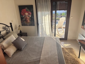 Quarto com cama, mesa de cabeceira com abajur, pintura e vista para a varanda.