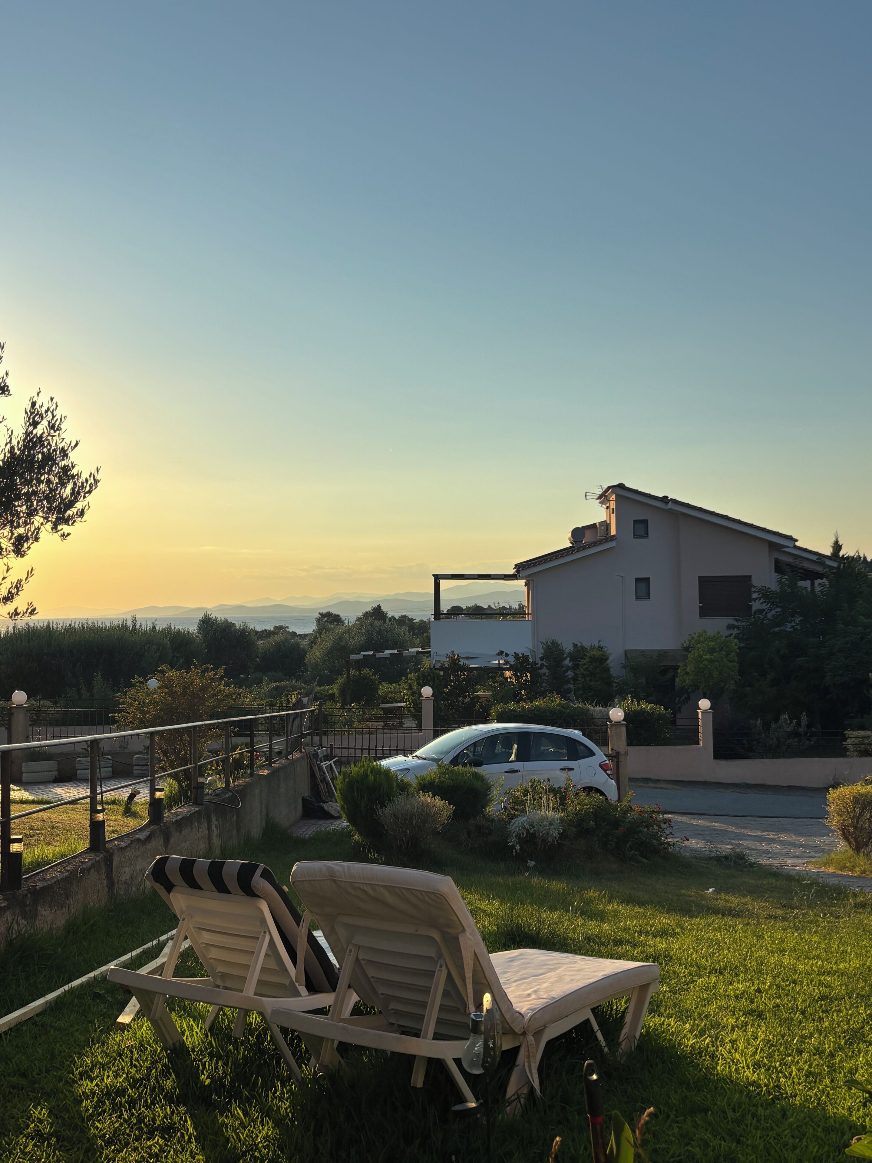 Ein modernes Haus mit einem geparkten Auto davor, einem Poolbereich mit Liegestühlen und freiem Blick auf die Landschaft.