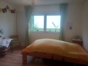 Ein Schlafzimmer mit einem Bett, einem Sessel, einem Tisch mit Tischdecke, einem Fenster mit Vorhängen und einer Wandlampe.