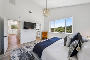 Un dormitorio moderno con una cama grande, televisor, vista a áreas verdes y una cocina al fondo.