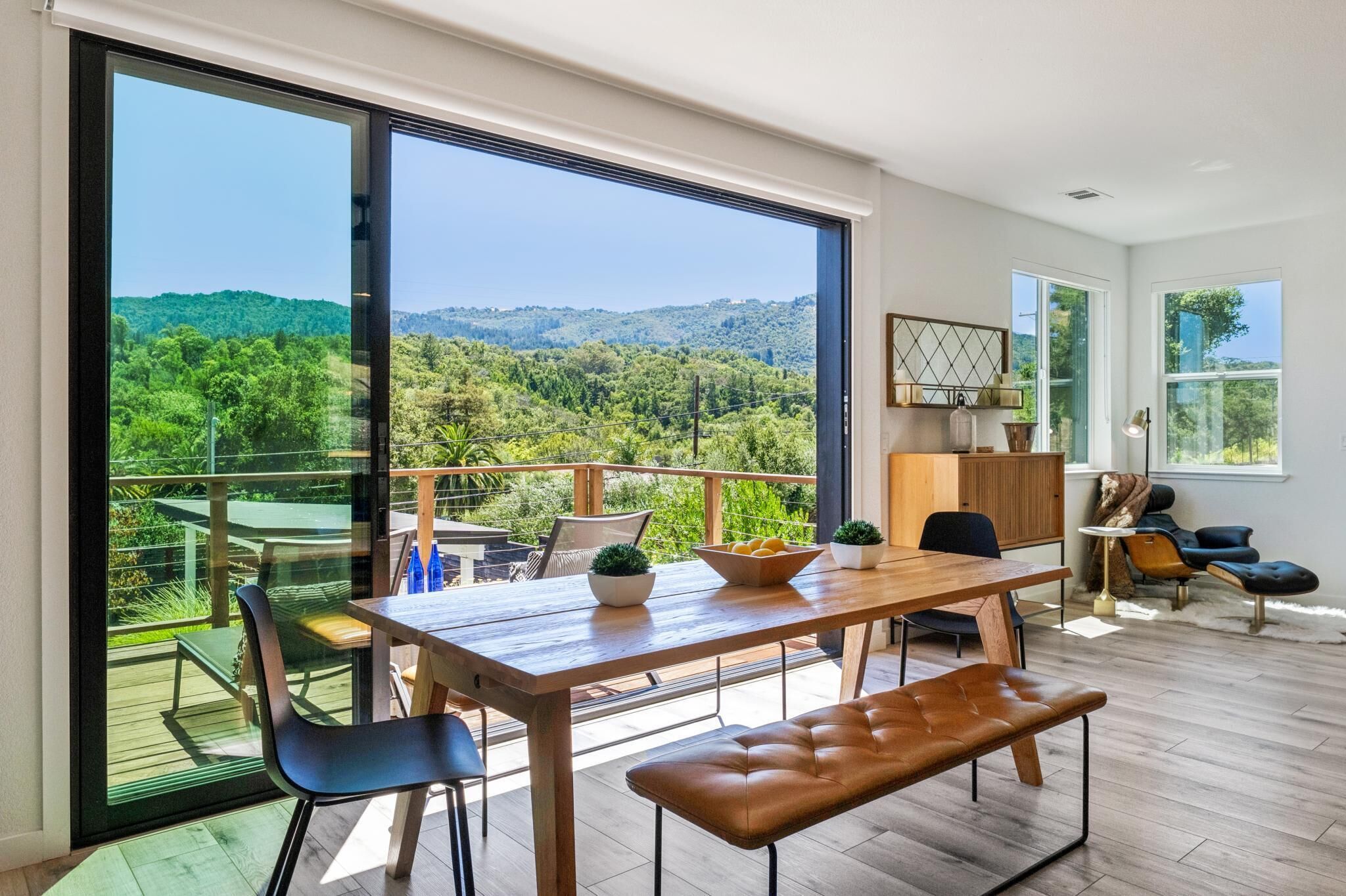 A spacious balcony with a wooden table and chairs, a bench, and a view of a lush green landscape.