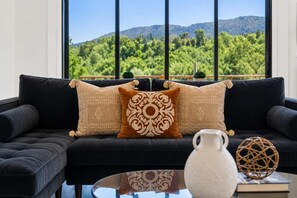 Un sofá negro con cojines estampados y un jarrón blanco sobre una mesa de vidrio, con vistas a la vegetación y montañas a través de amplios ventanales.