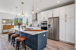 Una cocina moderna con armarios blancos, una isla grande con taburetes, electrodomésticos de acero inoxidable e iluminación colgante.