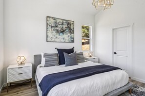 A bedroom with a large bed, two bedside tables, a nightstand, a geometric pendant light, a wall-mounted abstract painting, and a door. - The Vintara - Glen Ellen,Hot Tub, New Construction (Glen Ellen)